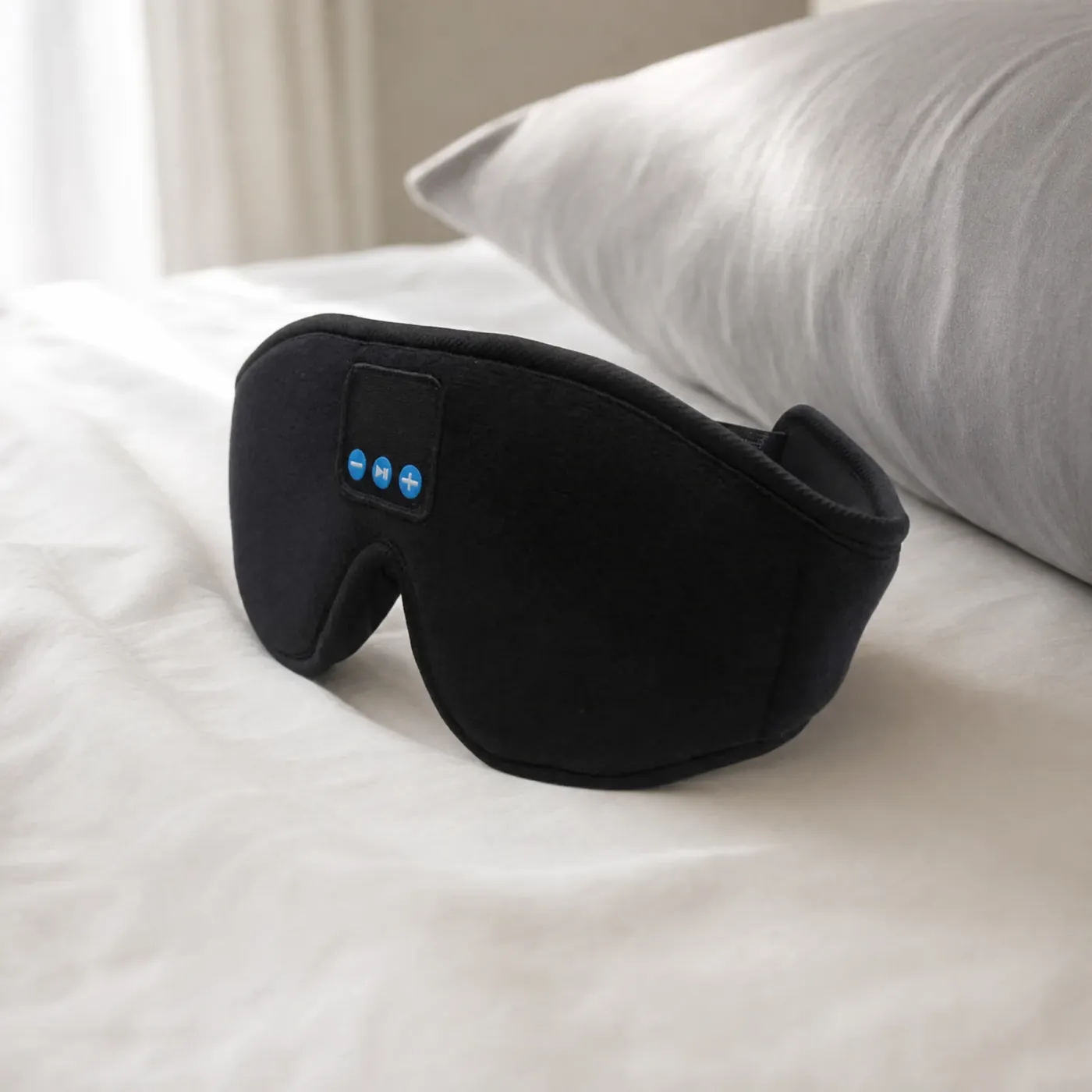 Black Bluetooth sleep mask with three control buttons on the front, lying on a white bed beside a grey pillow