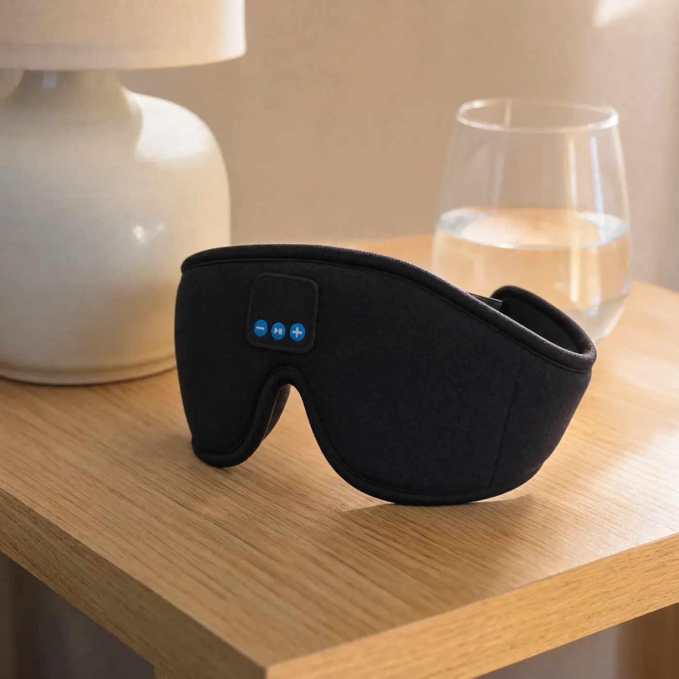 Black Bluetooth sleep mask on a wooden bedside table next to a lamp and a glass of water